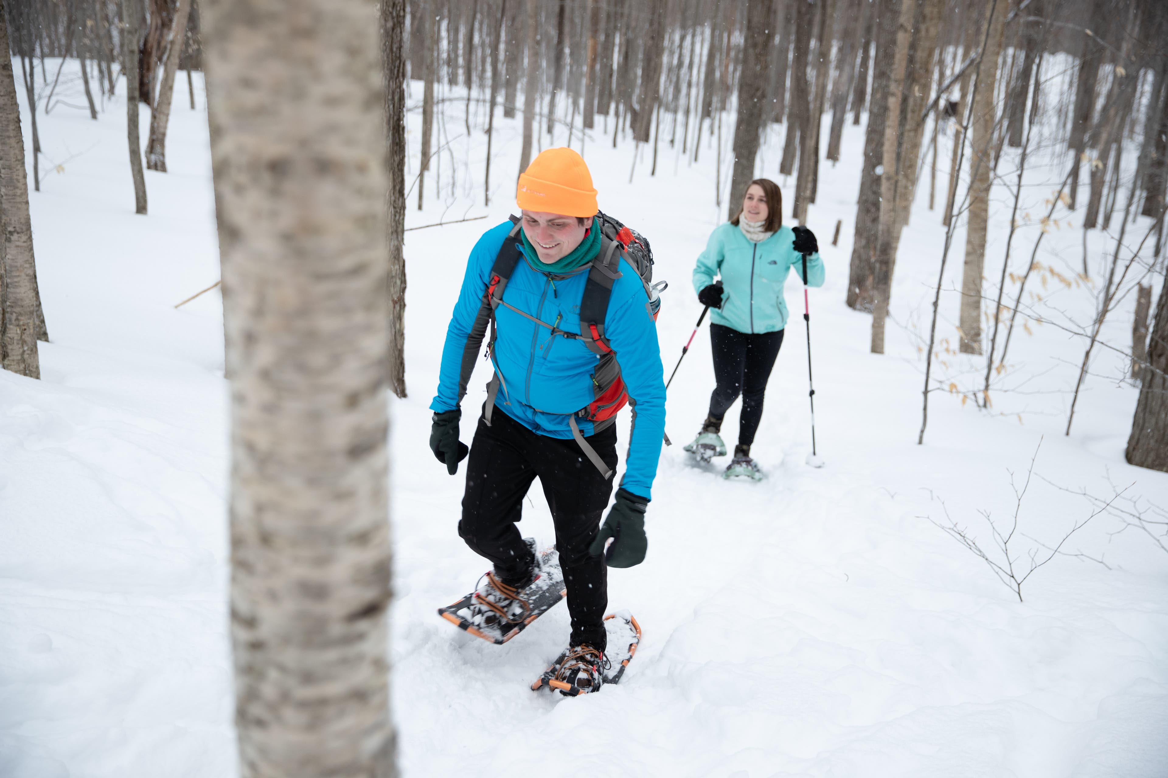 A white man and white woman wear blue and turquoise jackets and black pants as they snowshoe through the bare trees of the Adirondack Mountains.