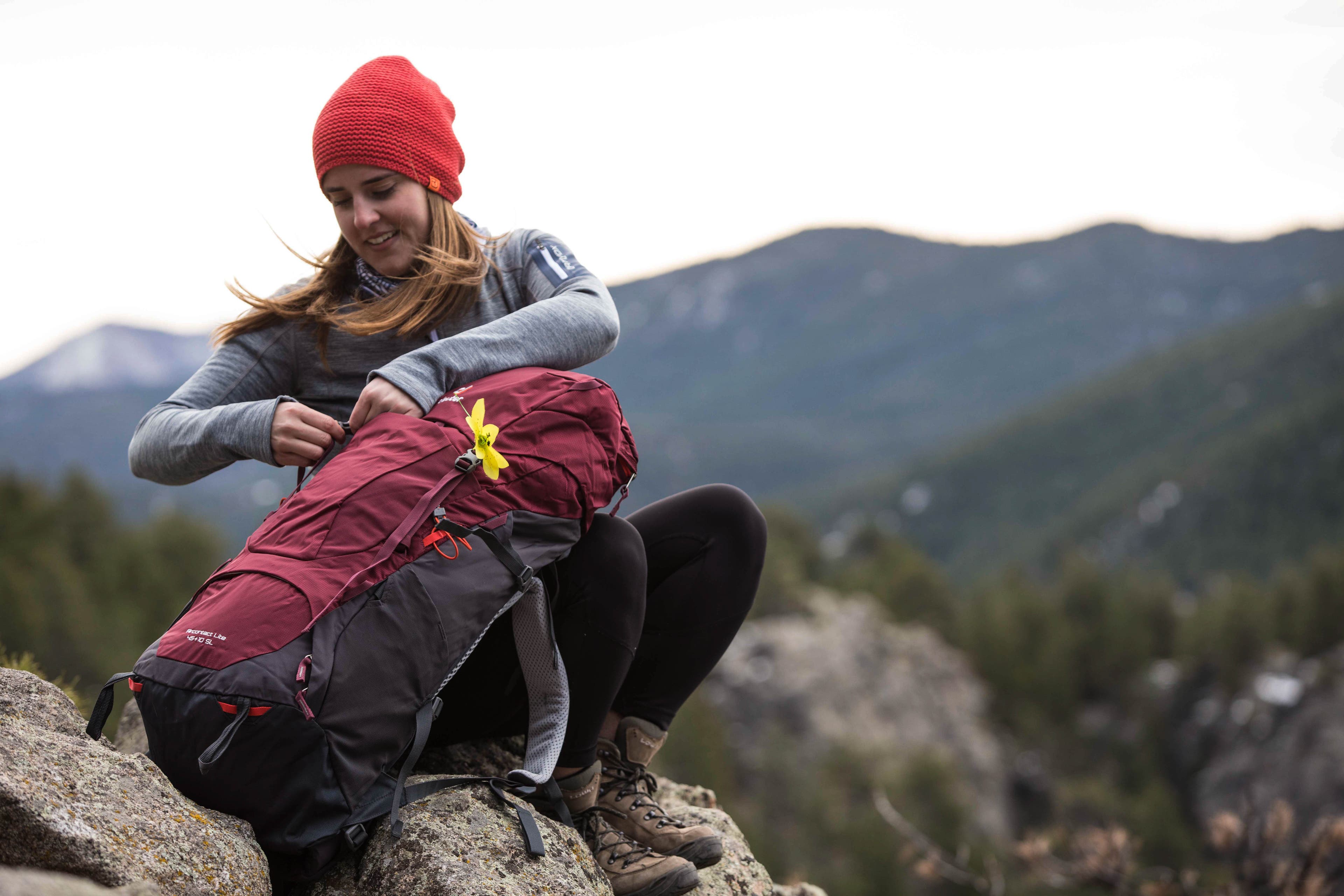 Aircontact Lite 45+10 SL Maroon Graphite-9 White woman sits on a rock holding a red Deuter Aircontact Lite backpack. Mountains are out of focus in the background.
