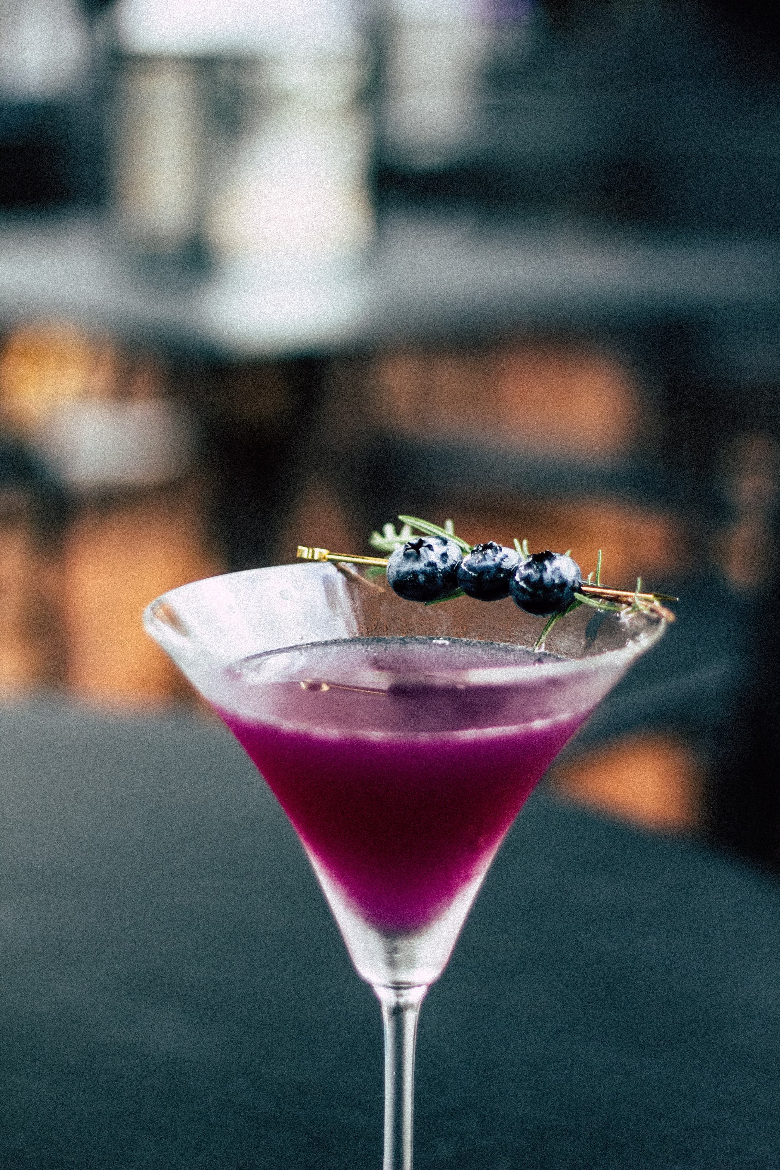 mgg-vitchakorn-T5T1mPwYAGA-unsplash Blueberry Pine Martini is in focus with a blurred background. Liquid is purple. A toothpick with blueberries and a sprig of pine needles lay across…