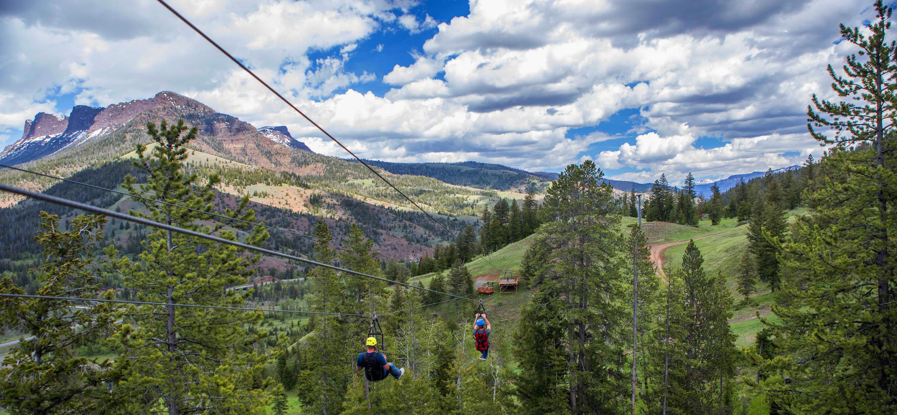 Two people zip-lining towards hill top with cloudy blue skies in background.