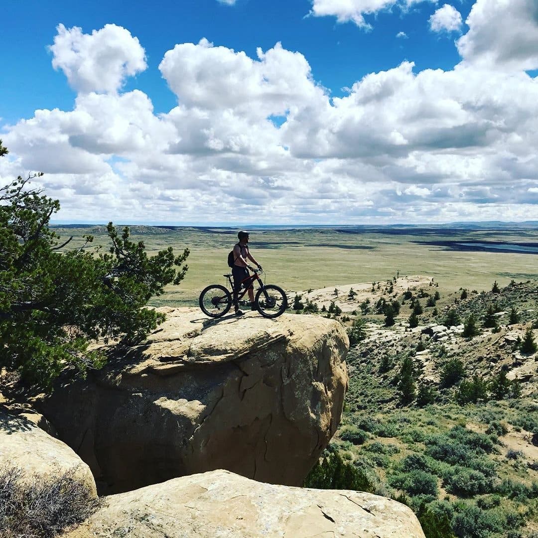 Man standing with mountain bike on slick rock with green fields and cloudy blue skies in background.