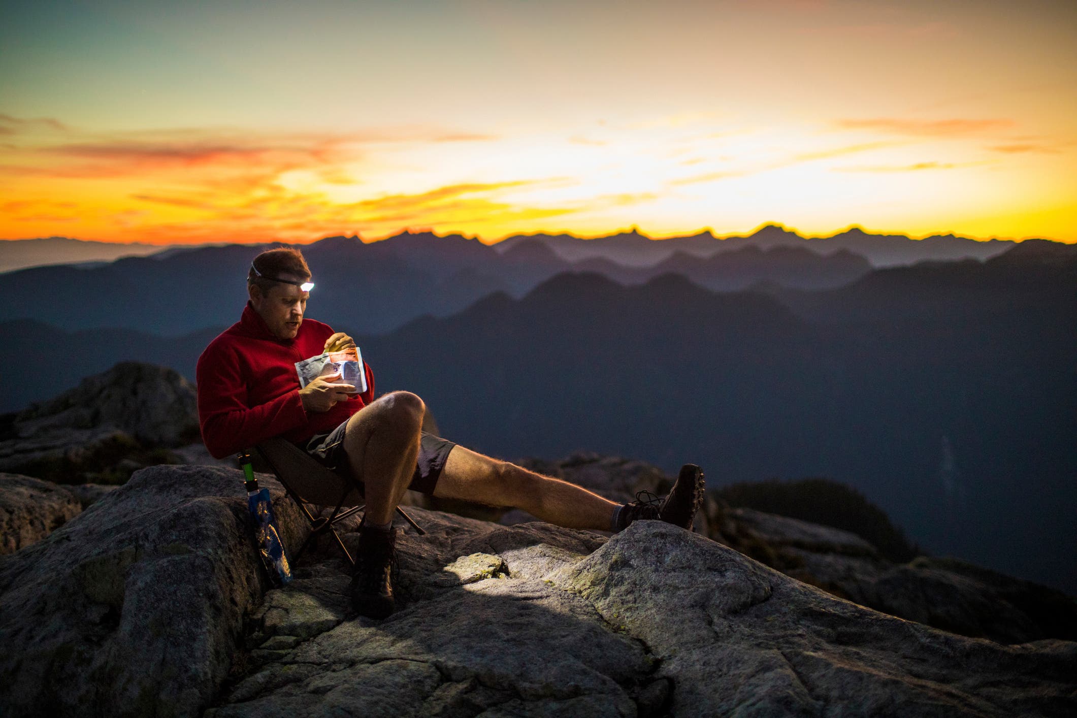 A backpacker relaxes on a summit, reclined in a camp chair. He is eating from a pouch of dehydrated food. Behind him is a colorful sunset.