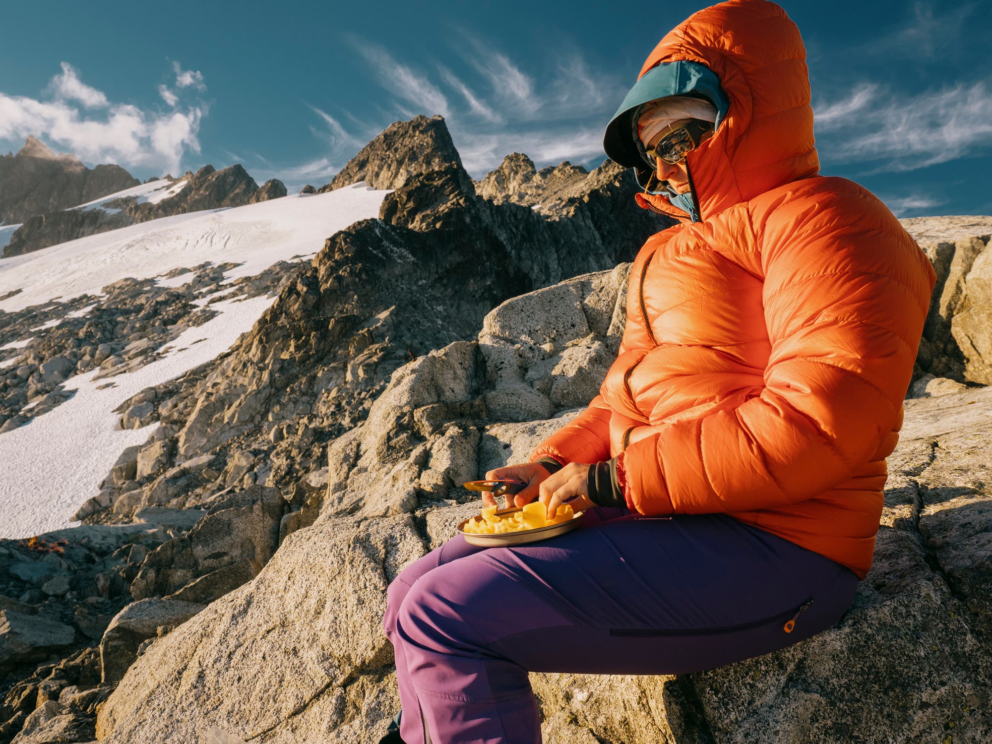 A hiker in a puffy down jacket sits on a rock with a plate of food in her lap. Mountains are in the background. 