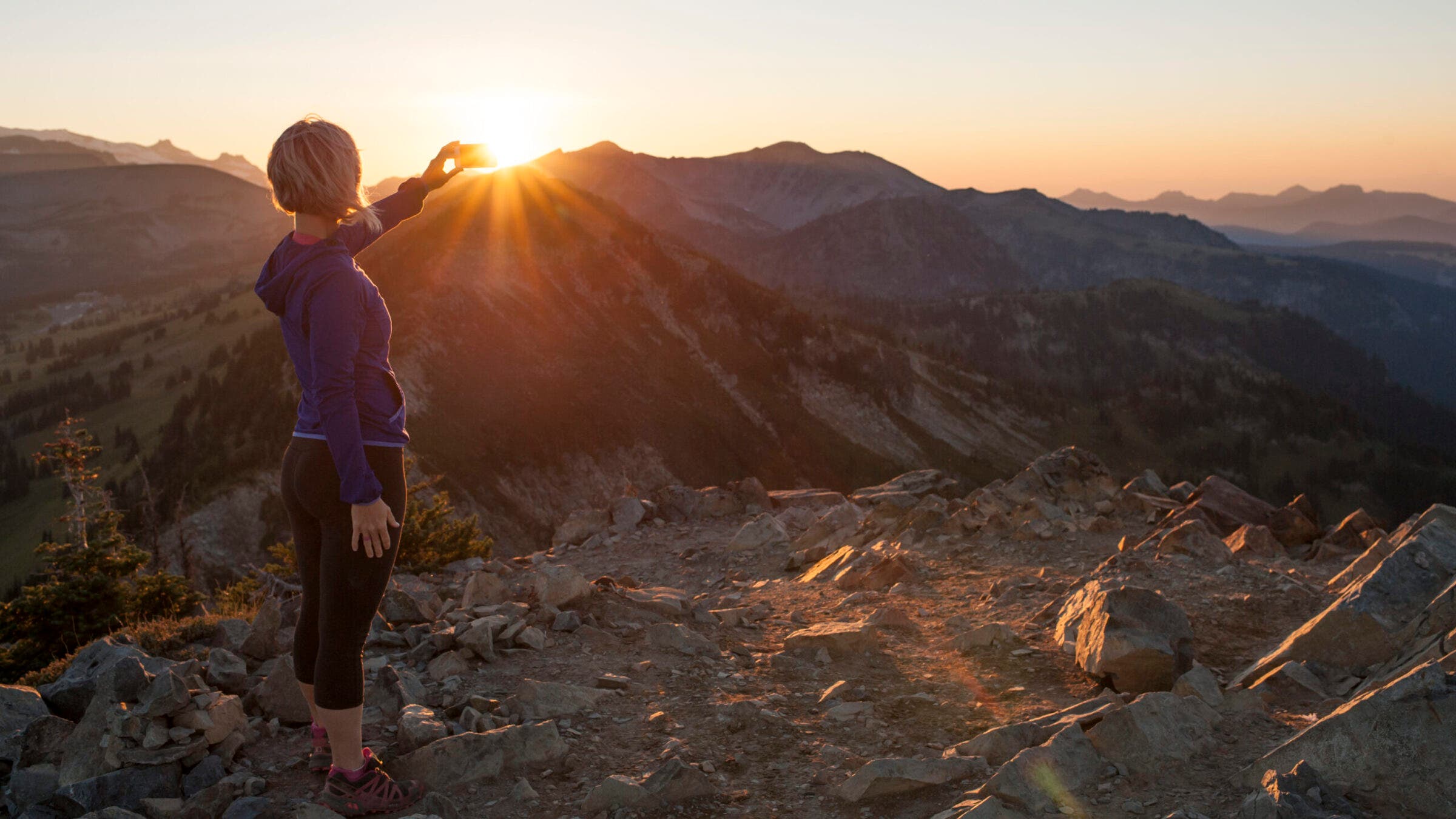 Hiker takes a photo of the sun peeking over the mountain after getting up early for one of the best sunrise hikes in the country. 