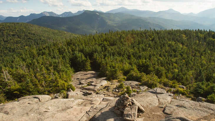 a rocky ridgeline trails away into thick woods with forested mountains rising beyond