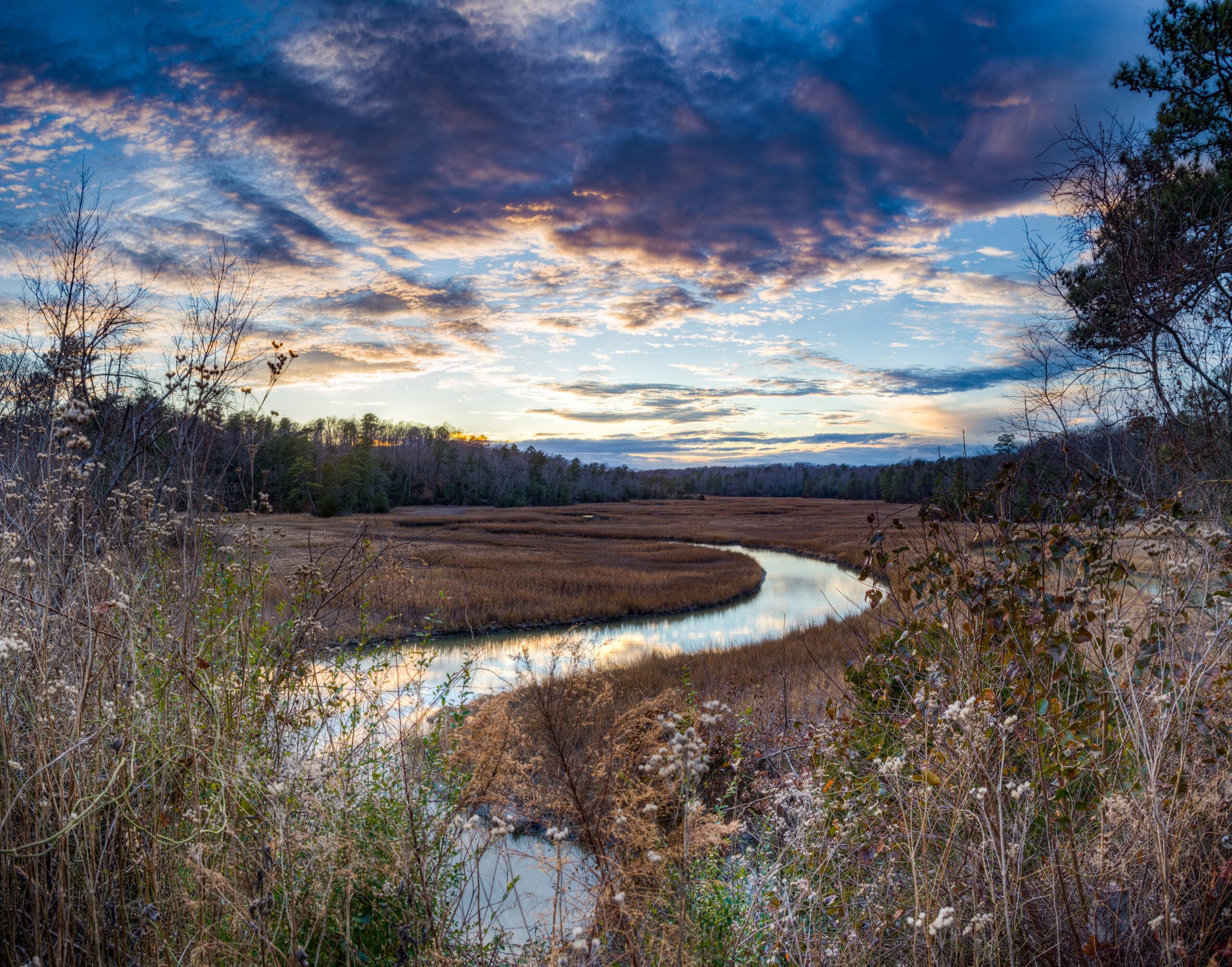 A river snakes a path through a field as sunset reflects off the water's surface. The deep blue sky is contrasted with bright highlights of orange as the sun lights the clouds from below.