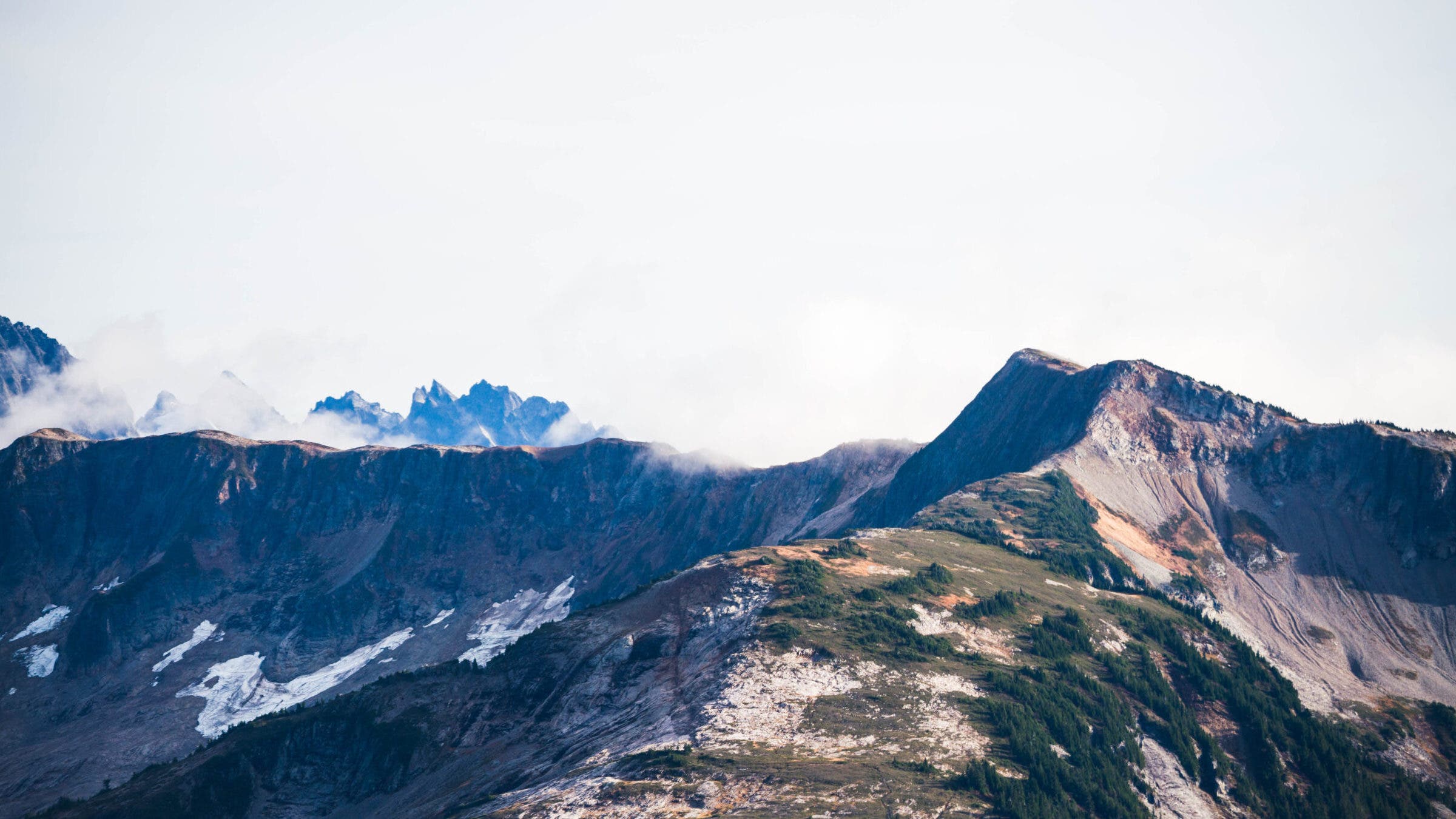 a broad ridgeline alternating dirt and meadow patches running into a background of spiky higher peaks