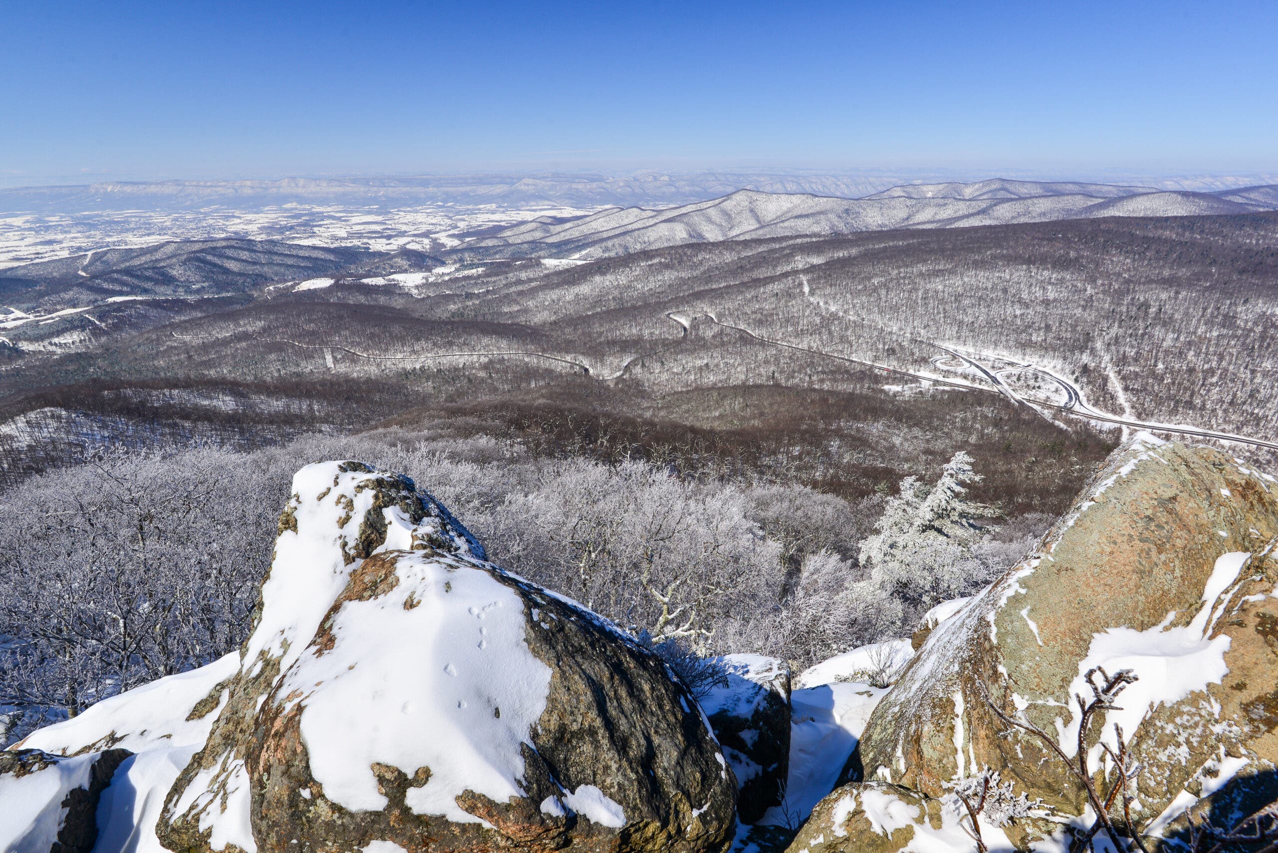 snowy boulders at the top of a mountain overlooking a valley full of snowy decidious forest with more mountains beyond
