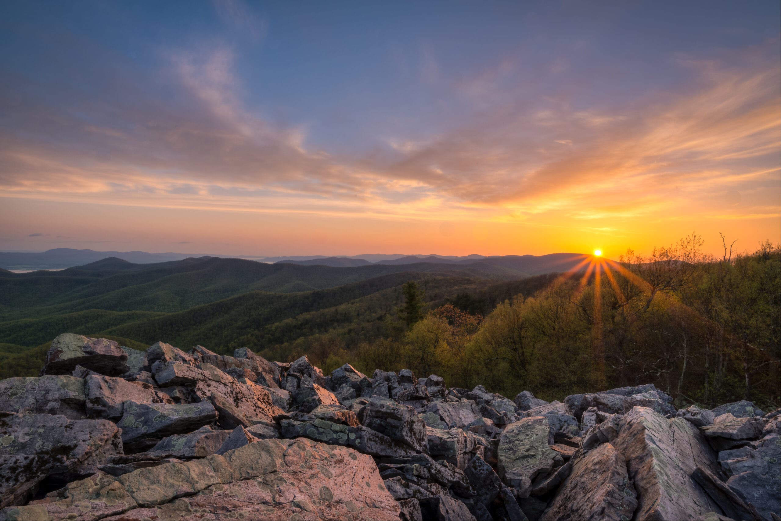a boulder field in the foreground with rolling, forested mountains beyond; the sun is coming up over the mountains