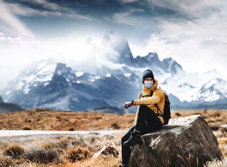 A hiker in a mask sits on a rock in front of a mountain vista. 