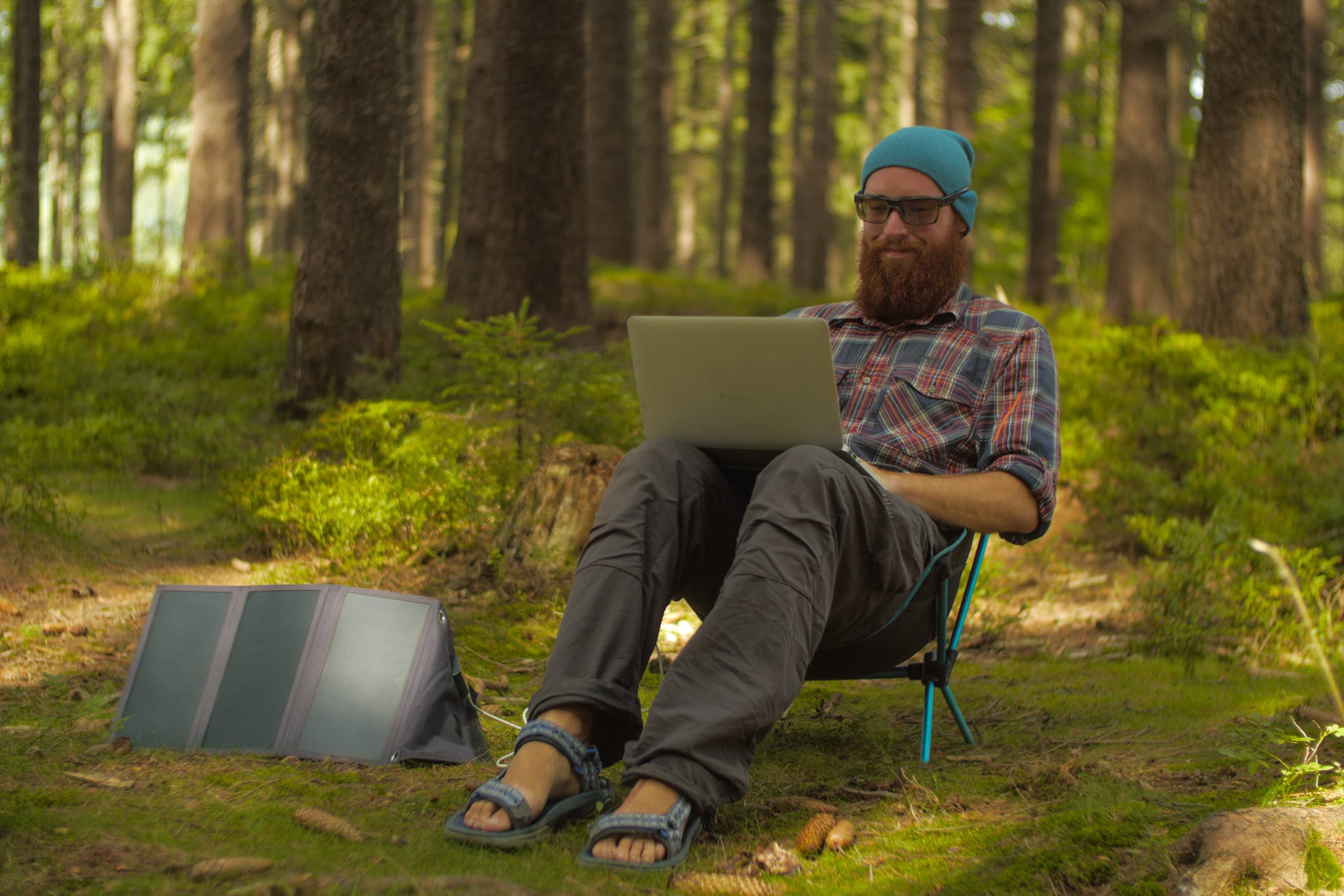 A man sitting in a camping chair in the forest working on a laptop. There's a solar panel on the ground next to him.