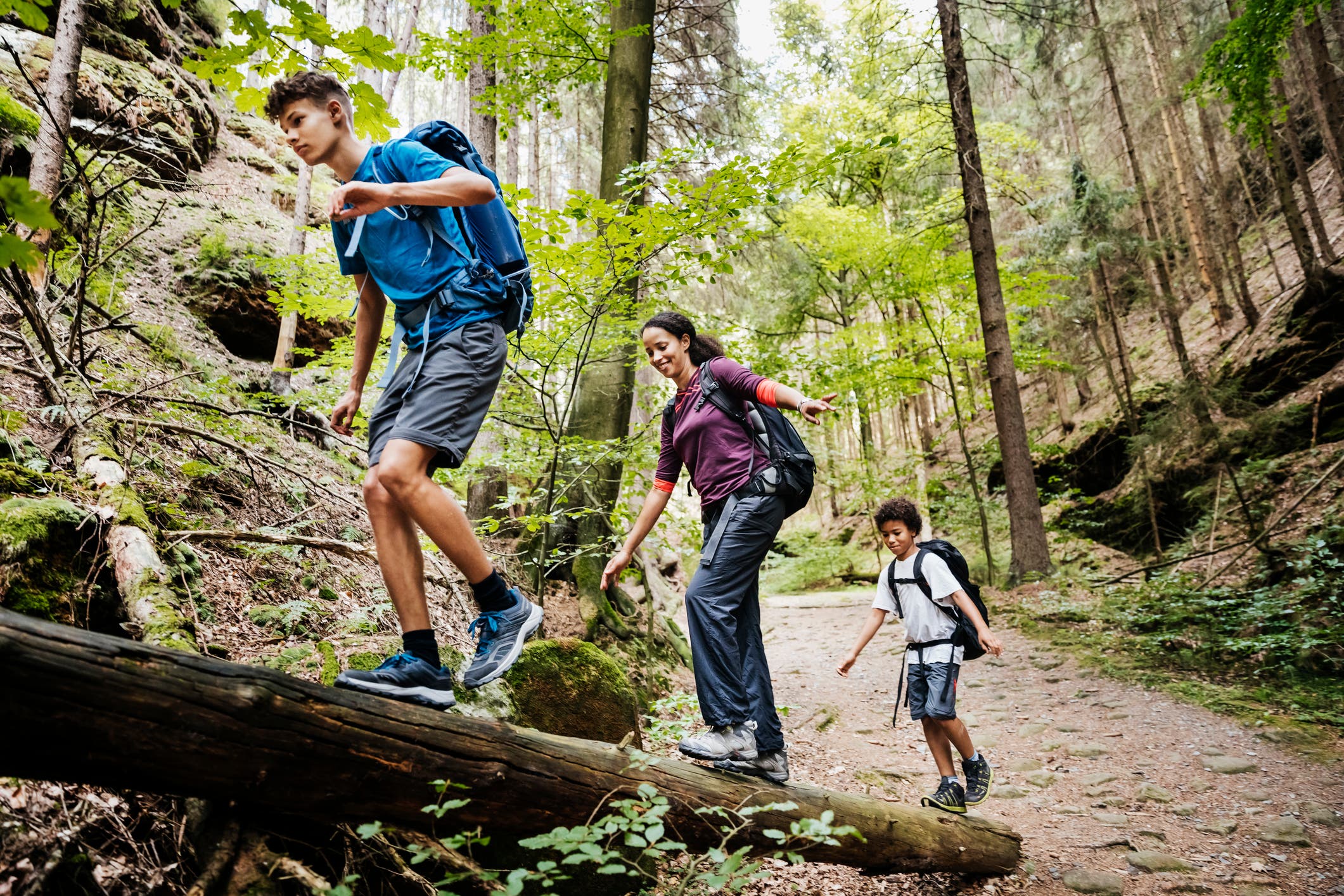 A family of three (a teenage boy, a mother, and young boy) hiking together. They are in a row walking over a fallen log. All are smiling and wearing backpacks. They're surrounded by trees.