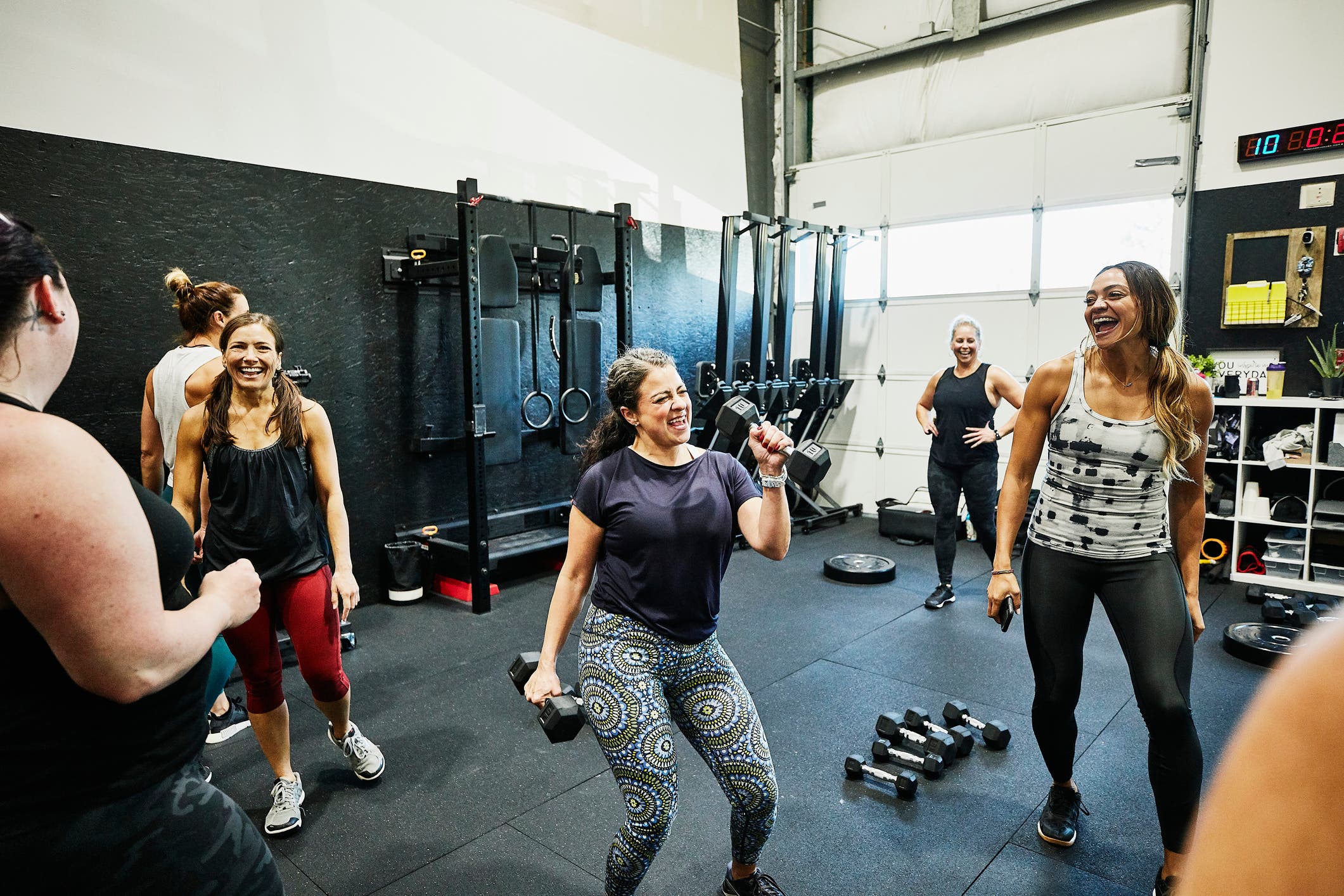 A woman in a gym smiles while holding two dumbbells. She's surrounded by other women in workout clothes. All are smiling. 