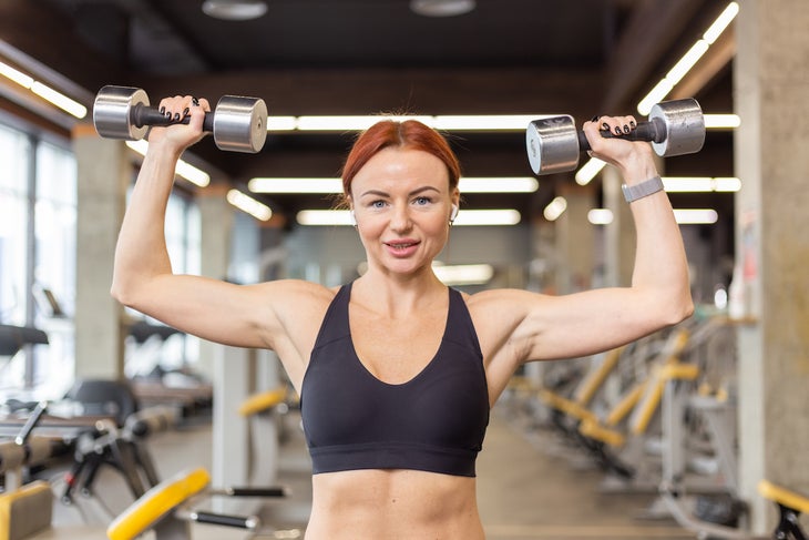 Woman trains shoulder muscles doing overhead dumbbell press in a gym.