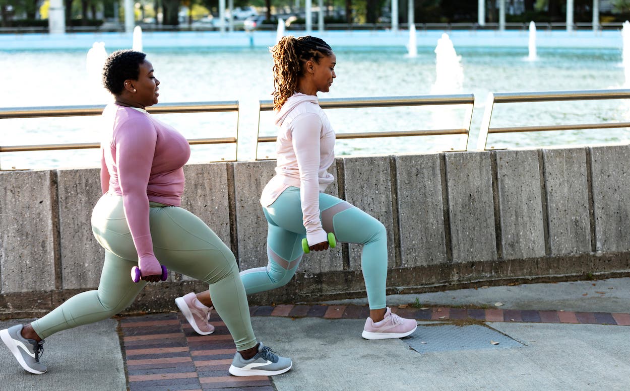 Two Black woman exercising together in a city park, by a large fountain on a city waterfront. They are doing lunges, carrying handweights.