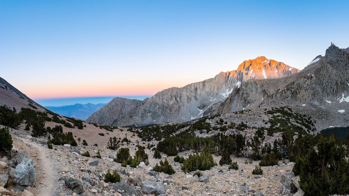 Rocky mountain path is illuminated by last rays of sunlight. It is a stark landscape with scattered trees, gray rock, and a small lake