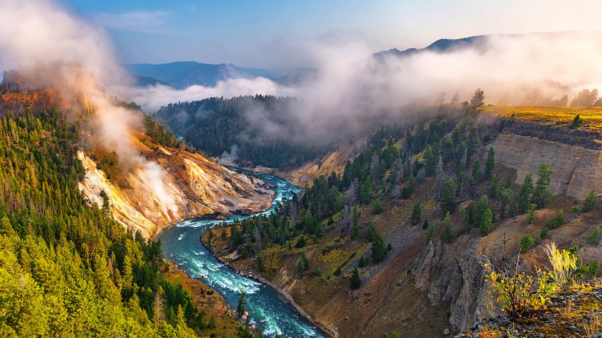 Calcite Springs View Area in Yellowstone National Park, Wyoming. Far below is the winding Yellowstone River, with the steaming Calcite Springs on the left bank of the river. Tall, tree-lined cliffs rise steeply on either side of the river and into the misty morning.