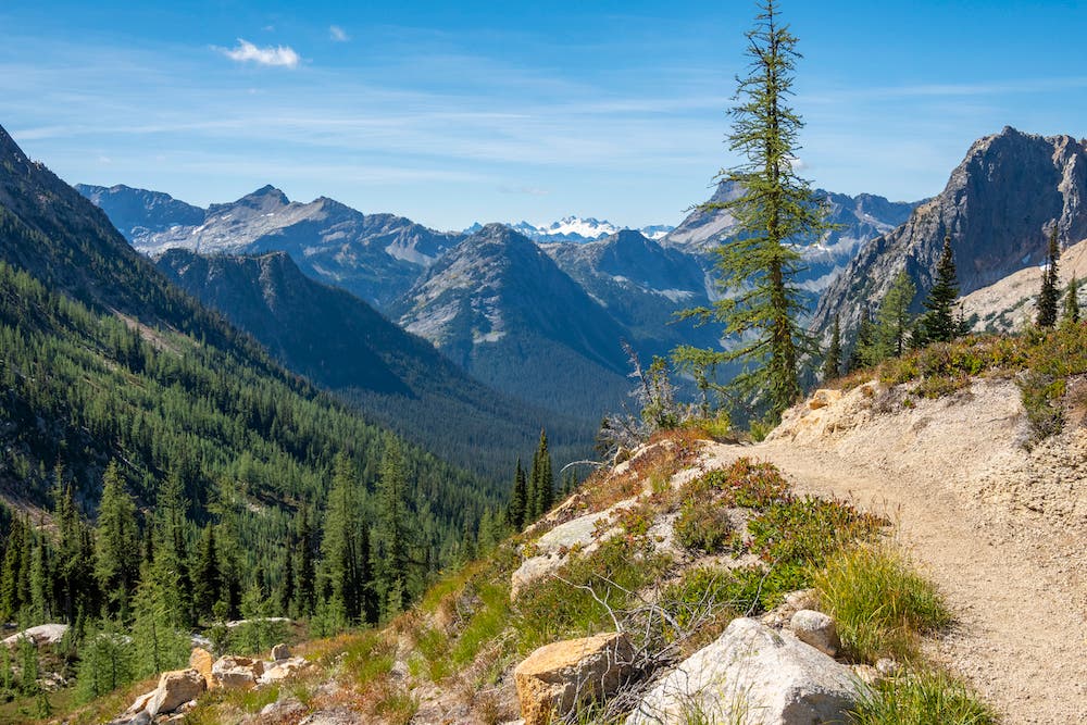 The Pacific Crest Trail winds around a high alpine ridge, with views of a deep valley and rugged North Cascades peaks in the distance.