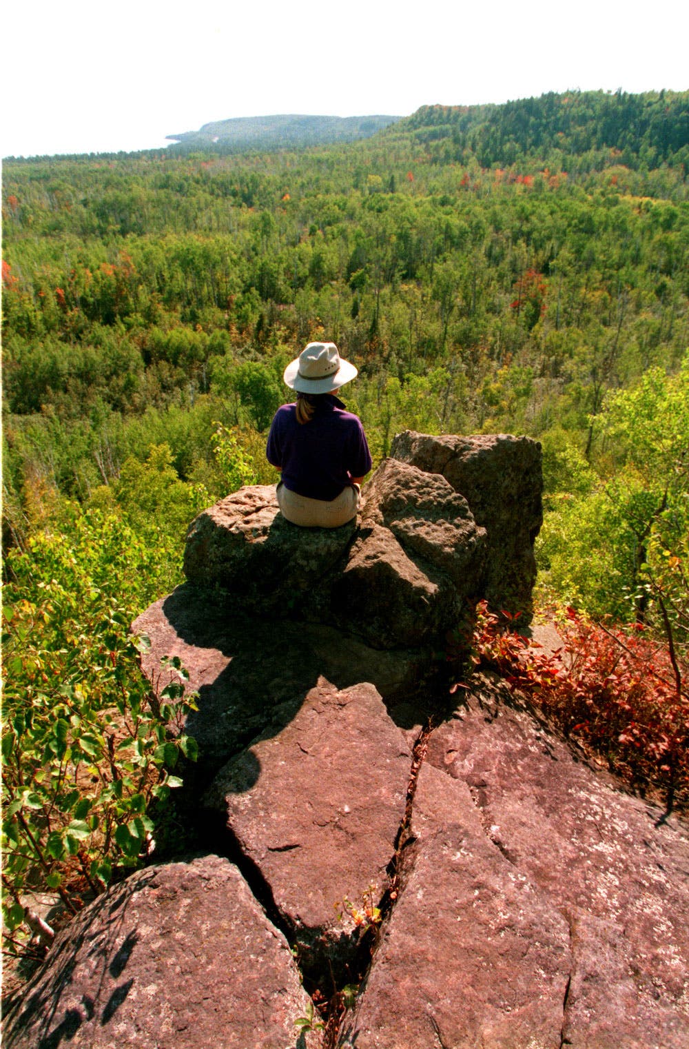 A HIKER SITS ON A LEDGE ALONG THE SUPERIOR HIKING TRAIL LOOKING TOWARD LAKE SUPERIOR