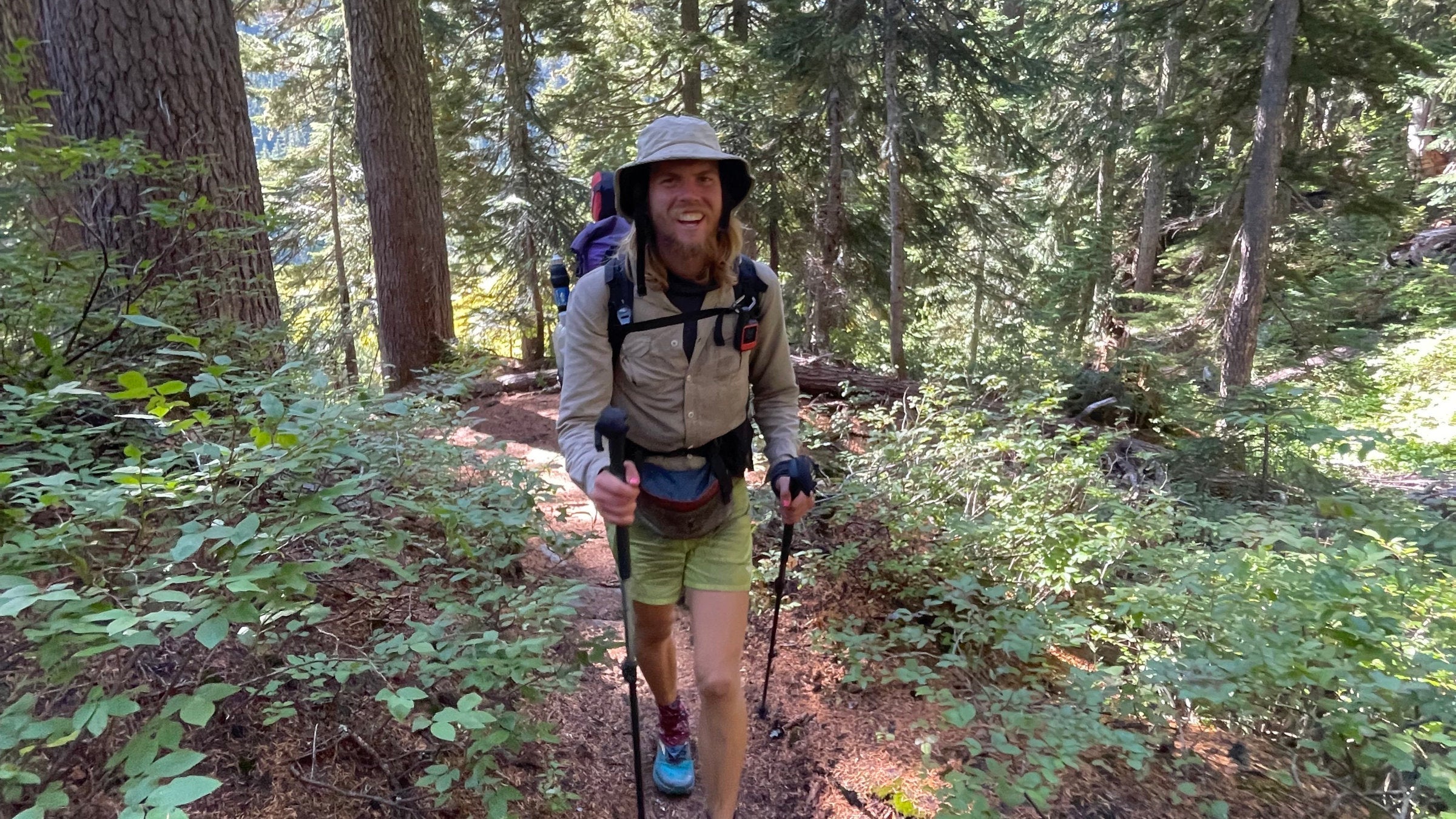 A hiker with trekking poles walks toward the camera smiling