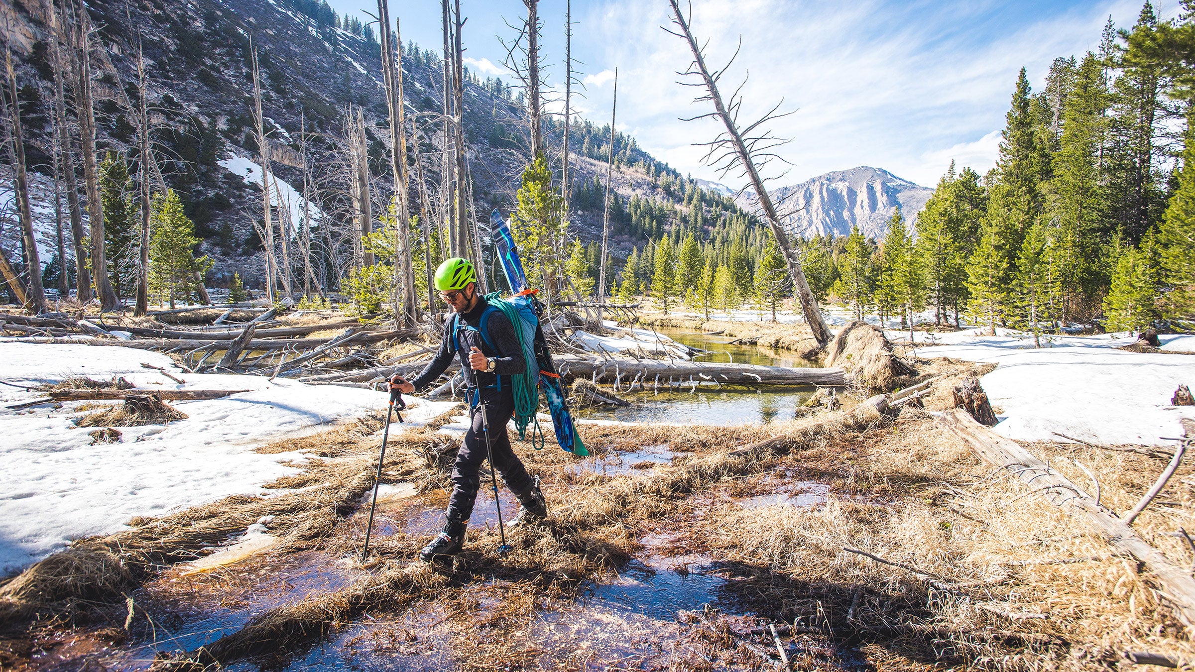 Man walking with skis on back across log in wooded area