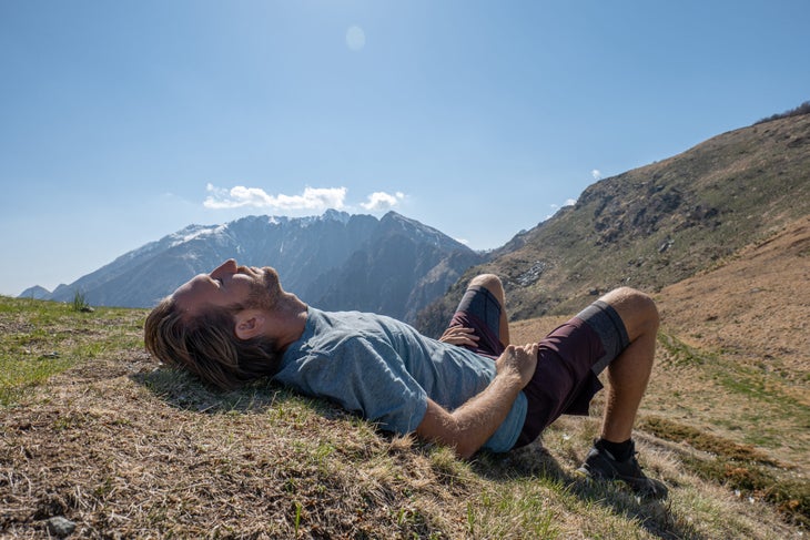 A man in shorts and a t-shirt lies on the ground in front of a mountain view. 