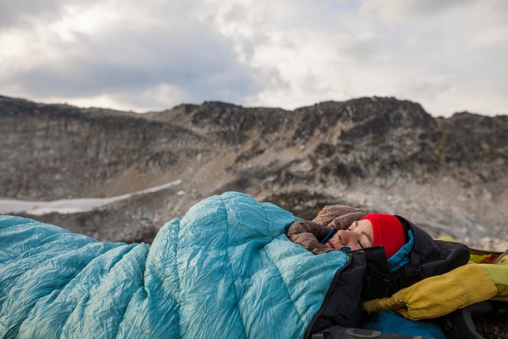 A hiker in a sleeping bag lies on the ground, with mountains in the background.