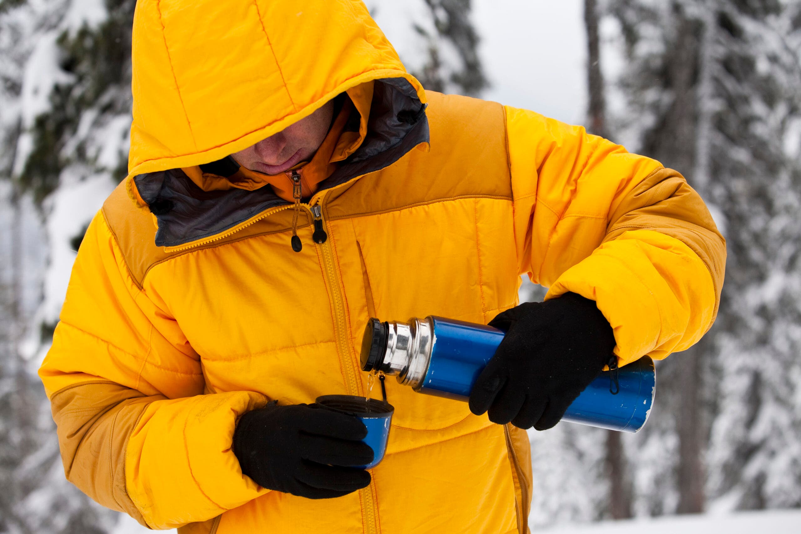 A man in a hooded yellow winter jacket and gloves pours hot liquid from a thermos.