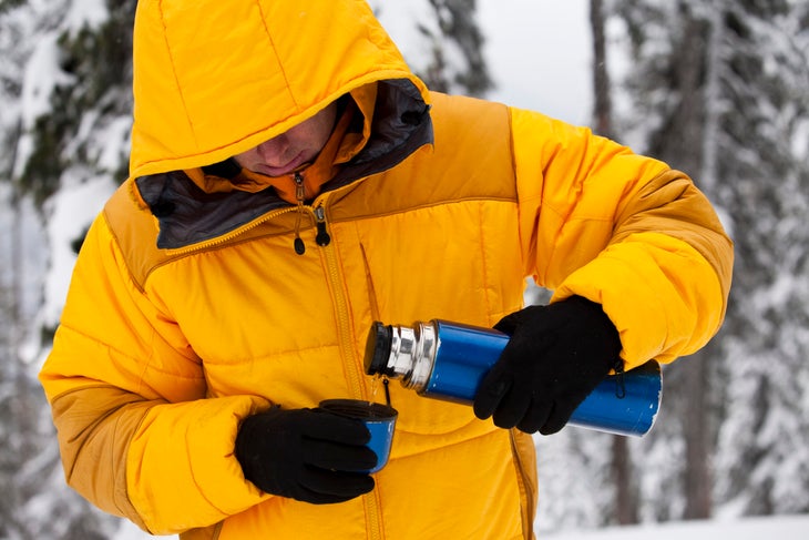 A man in a hooded yellow winter jacket and gloves pours hot liquid from a thermos.