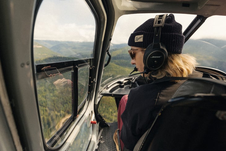 A woman in a beanie looks out the window of a helicopter at mountains