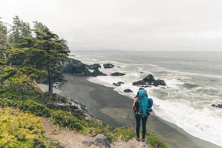 Female hiker with backpack standing on edge of cliff overlooking the pebble beach and ocean below on the west coast trail
