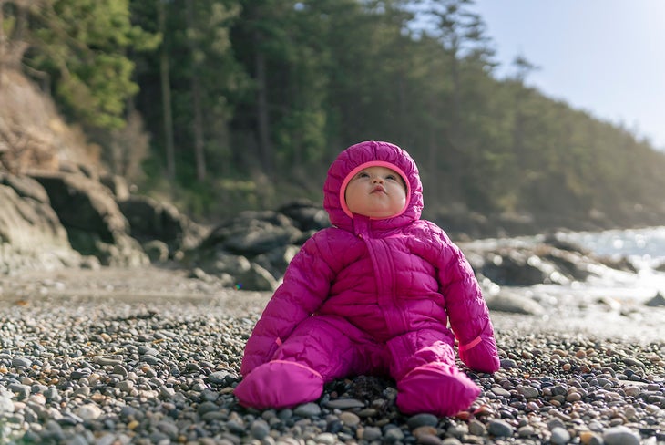 A young toddler is wearing a onesie snow suit and sitting on a bed of rocks at a Pacific Northwest beach. She is content and enjoying observing her surroundings.
