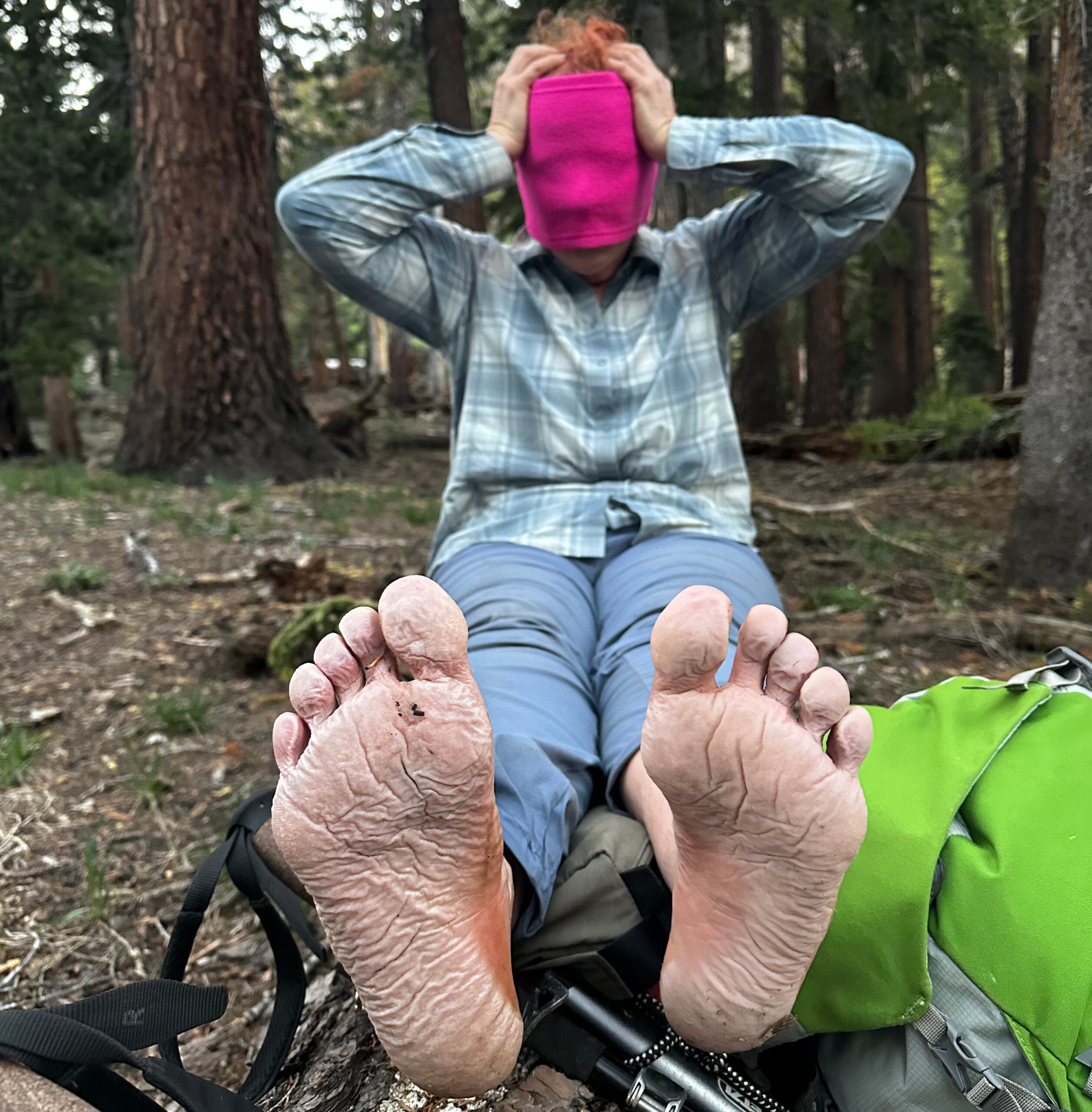 A female hiker with a neck gaiter pulled over her face shows her wrinkly feet to the camera