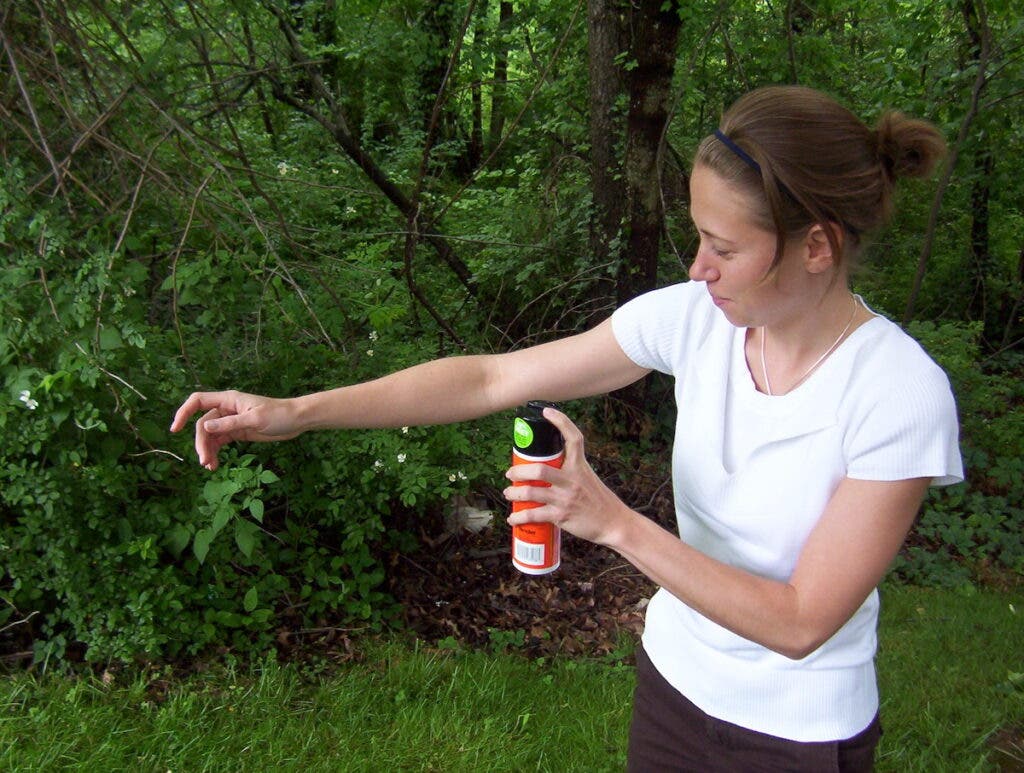 A woman sprays mosquito repellent on her arm in a shady forest.