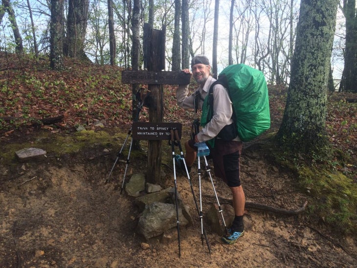 A thru-hiker poses beside a sign on a wooded area along the Appalachian Trail