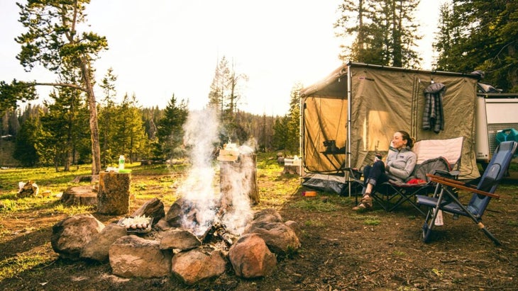 woman drinking coffee in a camp chair beside a campfire in the morning