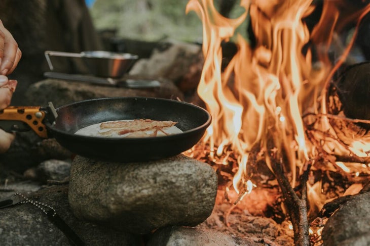 Eggs and bacon cooking in a skillet beside a campfire 