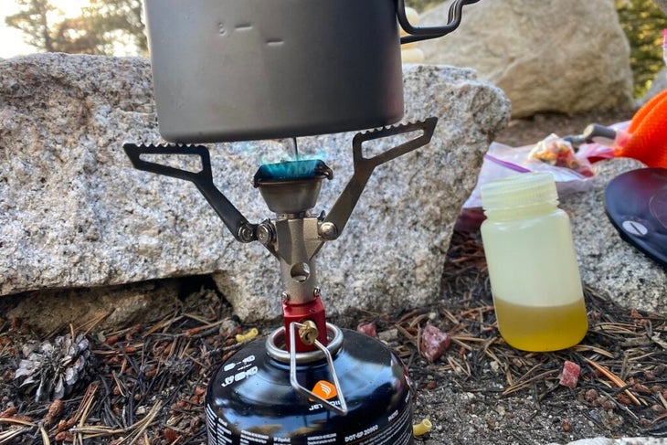 A lit MSR pocket rocket stove atop a black isobutane fuel canister outdoors near a rock. 