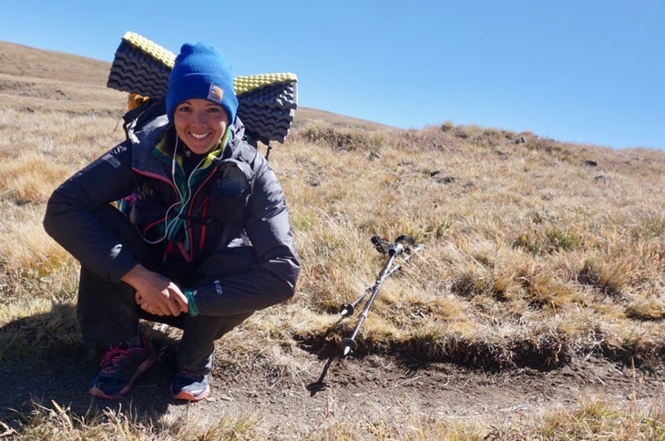 A backpacker wearing a blue beanie, jacket, and backpack sits on the ground beside a trail, smiling.