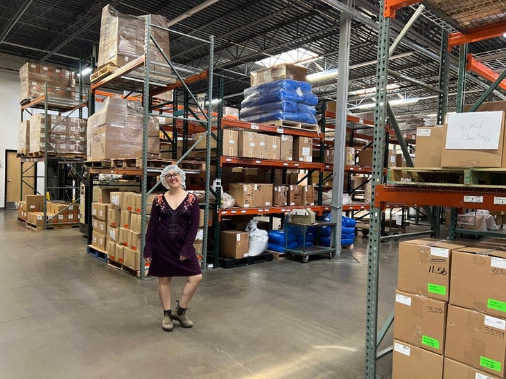 A woman in a dress and hairnet stands in a factory warehouse beside shelves of cardboard boxes.