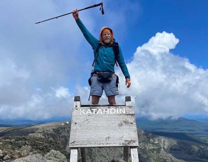 A thru-hiker stands on top of the Mount Katahdin sign on the Appalachian Trail on a sunny day.