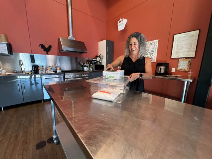 A woman stands in a kitchen with red walls, mixing ingredients at a long stainless steel table. 