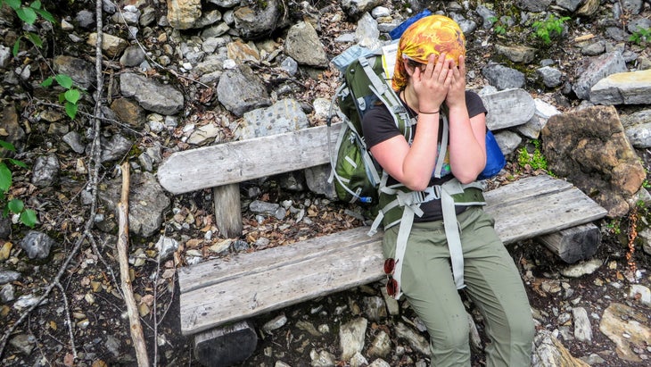 A close up of an exhausted female hiker sitting down on a bench with her hands over her face