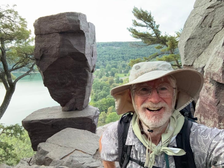 A hiker poses with a rock balancing on top of a boulder.