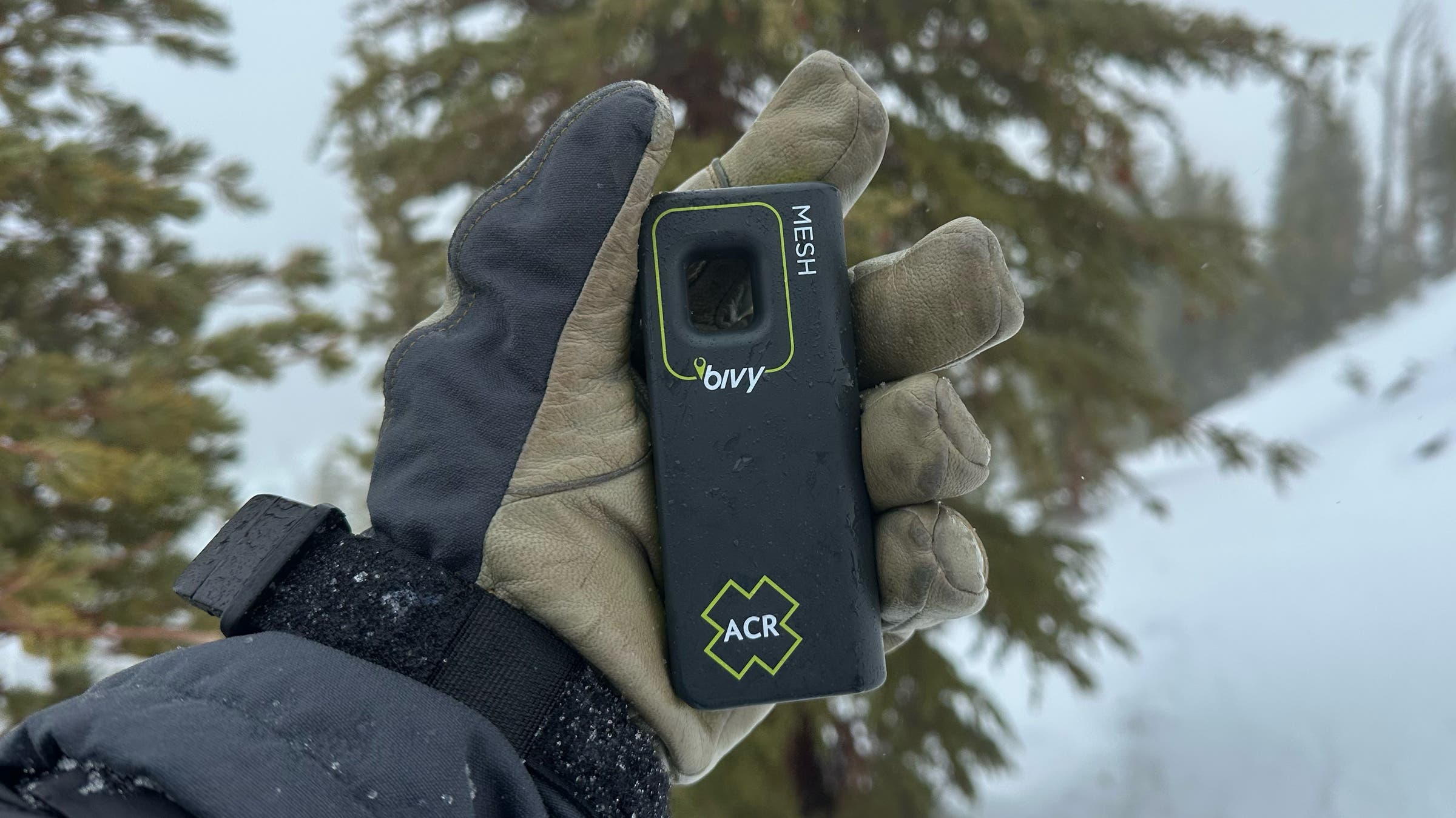 A person wearing a tactical outdoor glove holds the ACR Bivy Stick Mesh, the best satellite communicator for groups, against a blurry backdrop of snow-dusted evergreen trees.