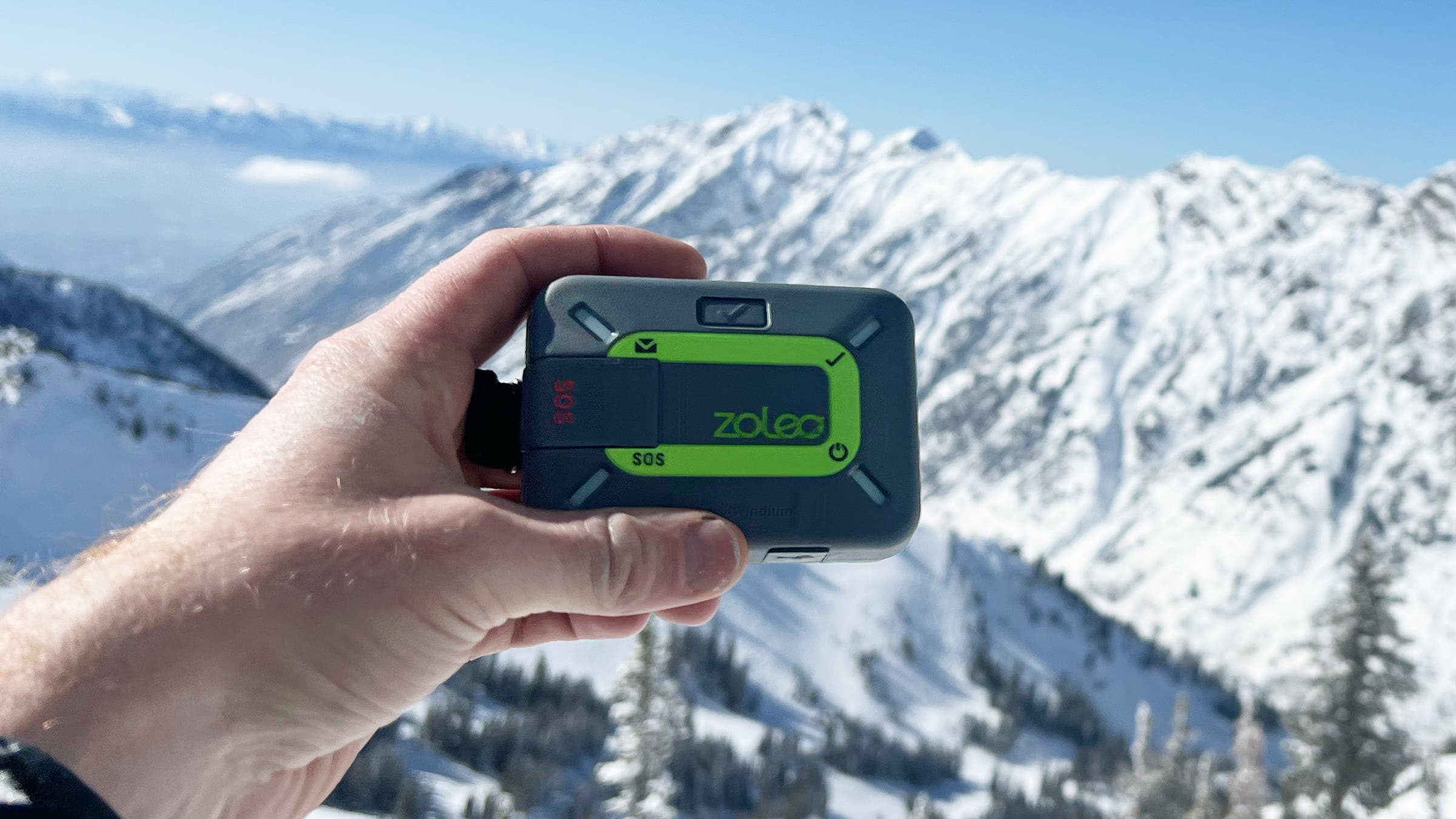 The Zoleo Satellite Communicator, the best budget satellite communicator, is held against a background of snow-covered mountain peaks, showcasing its rugged, portable design for affordable off-grid safety.