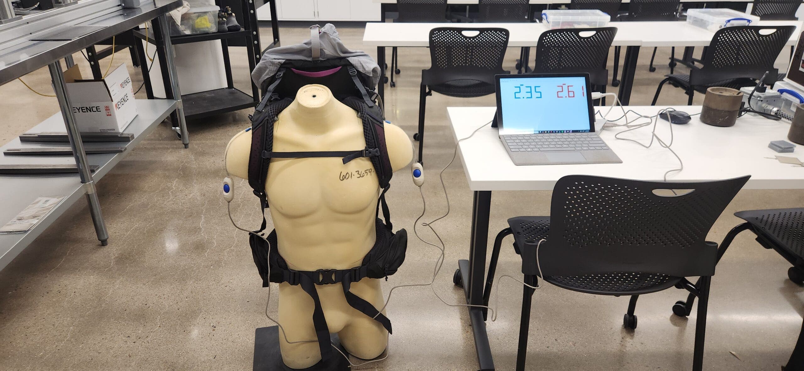 A tan male torso mannequin wears a technical backpack equipped with electronic sensors on the shoulder straps and hip belt for backpack load testing. The sensors are wired to a nearby laptop displaying real-time data, used to measure weight distribution between the shoulders and hips.