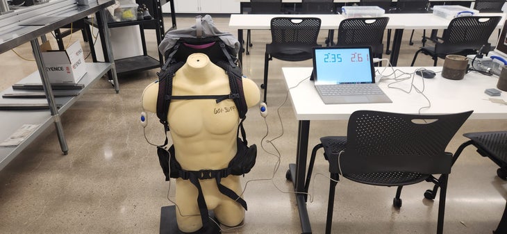 A tan male torso mannequin wears a technical backpack equipped with electronic sensors on the shoulder straps and hip belt for backpack load testing. The sensors are wired to a nearby laptop displaying real-time data, used to measure weight distribution between the shoulders and hips.