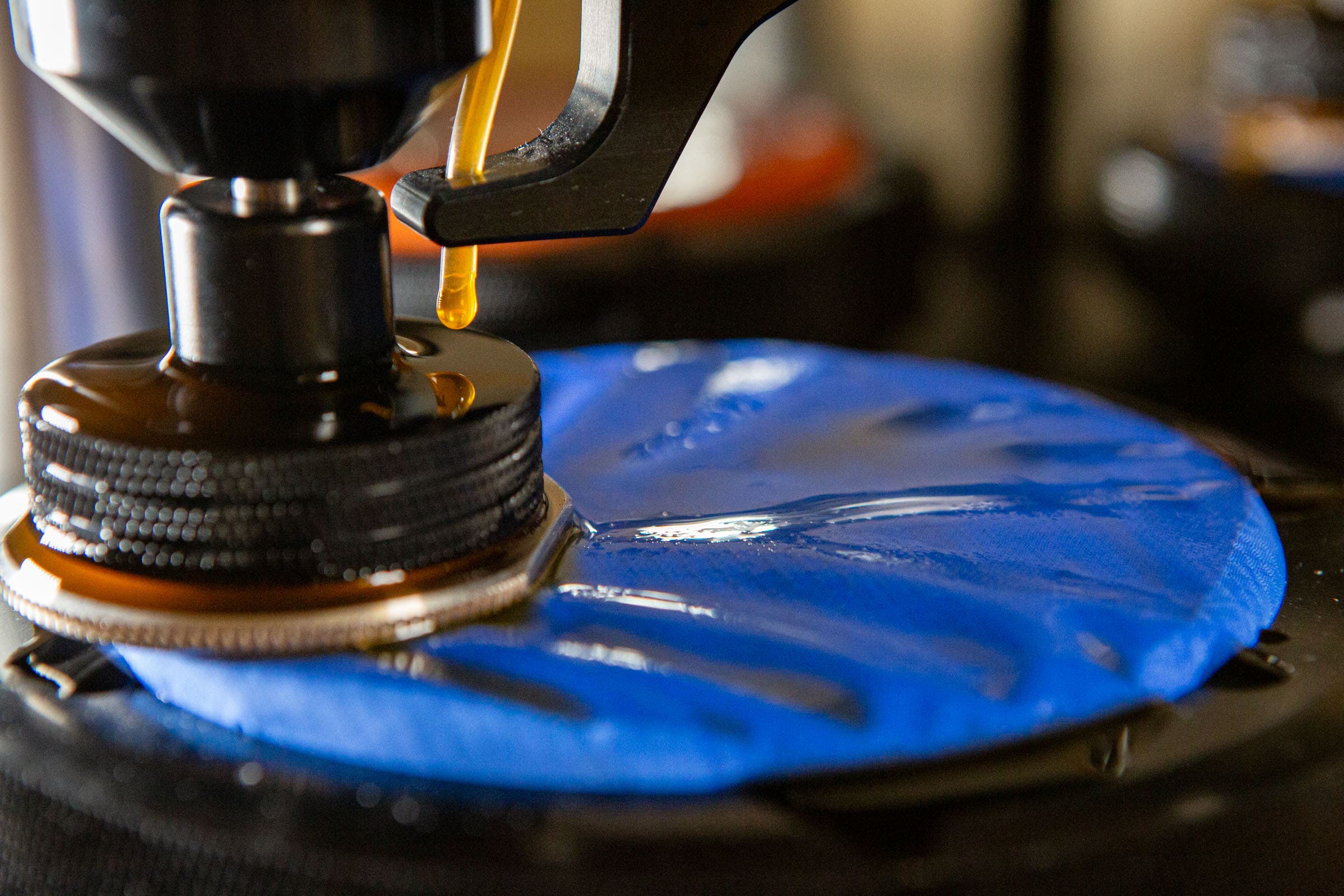 A detailed close-up of a blue fabric swatch undergoing rain jacket durability testing in a laboratory. A mechanical probe is applying pressure and a liquid solution to the wet material to measure how well the fabric resists wear and maintains waterproofness under friction.