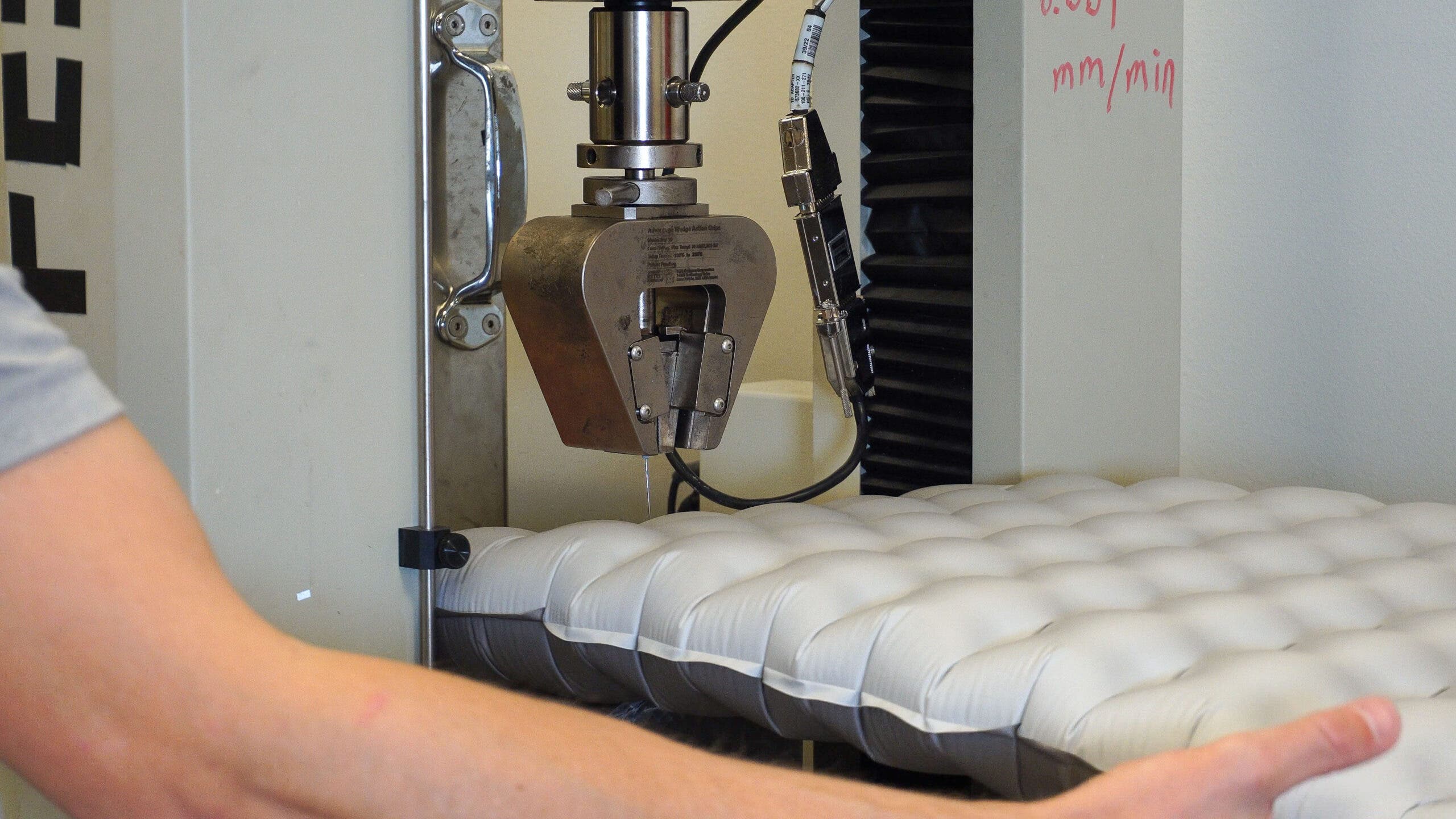 An inflated grey sleeping pad is positioned underneath a large industrial testing machine for a sleeping pad puncture test. A mechanical arm equipped with a fine needle is shown descending toward the pad's surface to measure the force required to pierce the material.